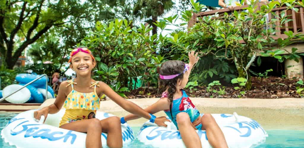 Two smiling girls in colorful swimsuits and goggles enjoy floating on inner tubes in a backyard pool with trees and a deck in the background