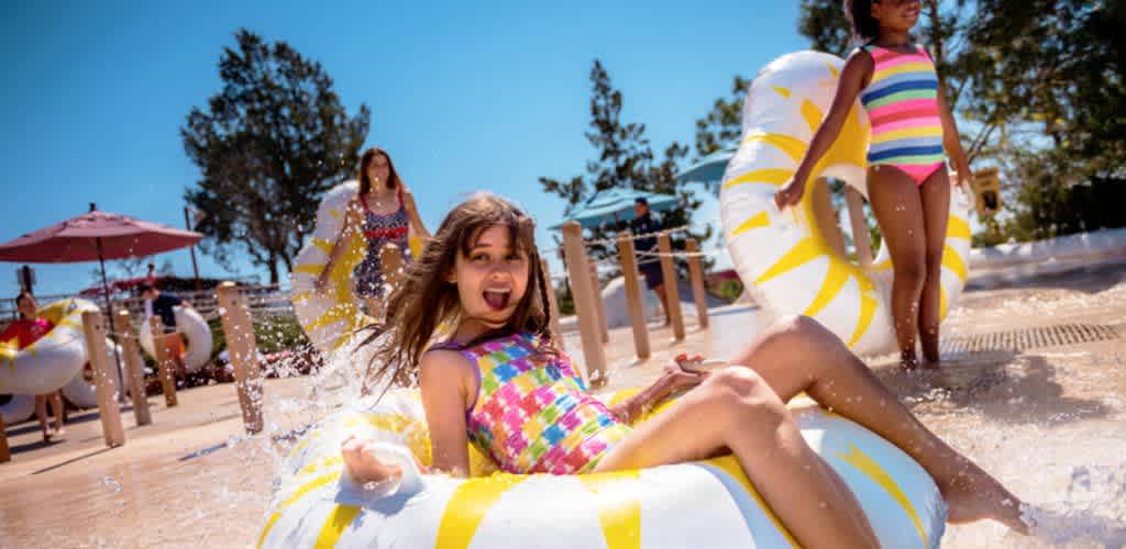 Three children in colorful swimsuits playing on pool floats and splashing water at a sunny outdoor water park