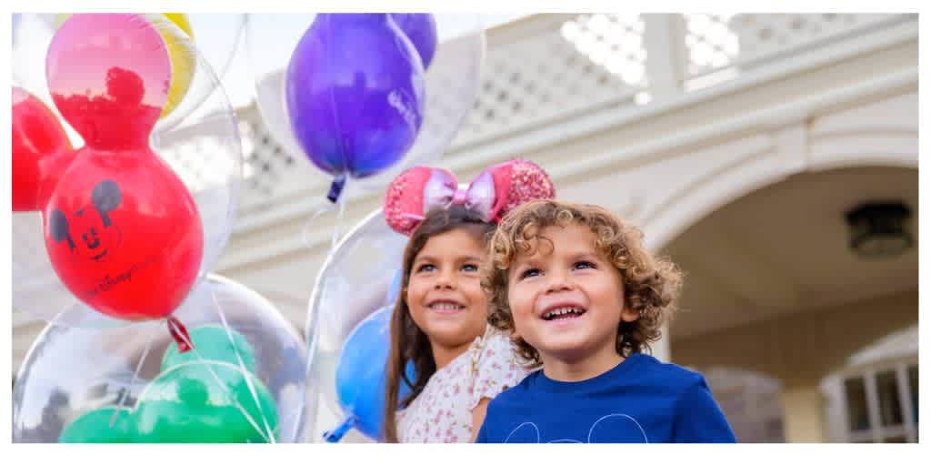 Two children smiling outdoors with colorful balloons in front of a house porch.