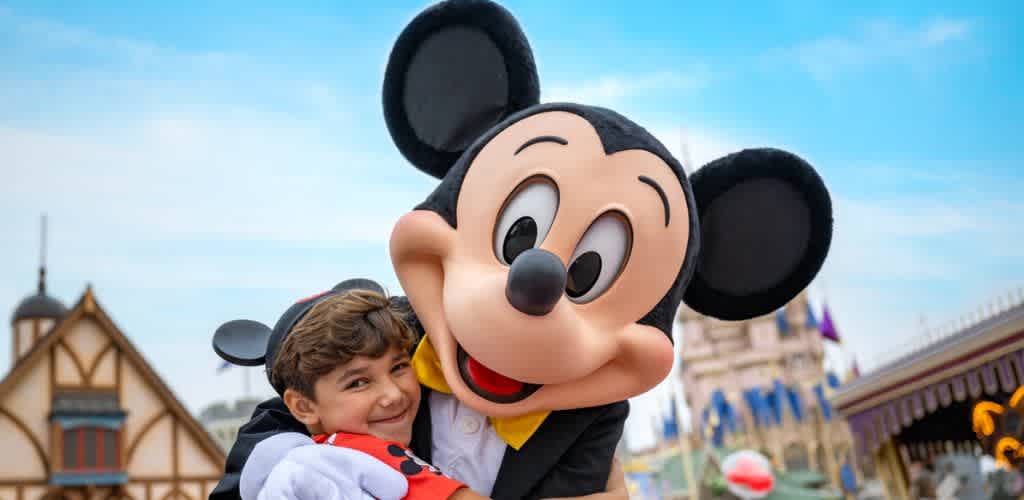 A young boy hugging Mickey Mouse mascot at a Disney theme park with buildings and a blue sky in the background