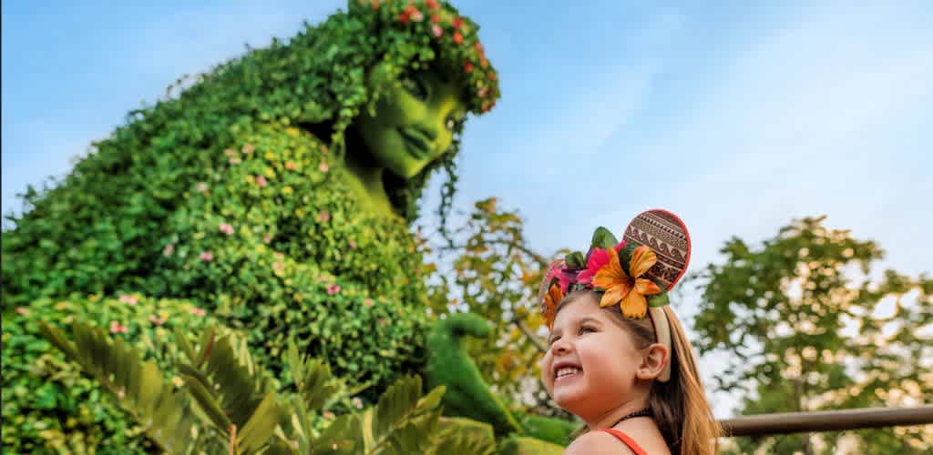 A girl wearing a colorful floral headband smiles at a large statue of a woman covered in greenery with flowers, set outdoors against a bright sky and trees.