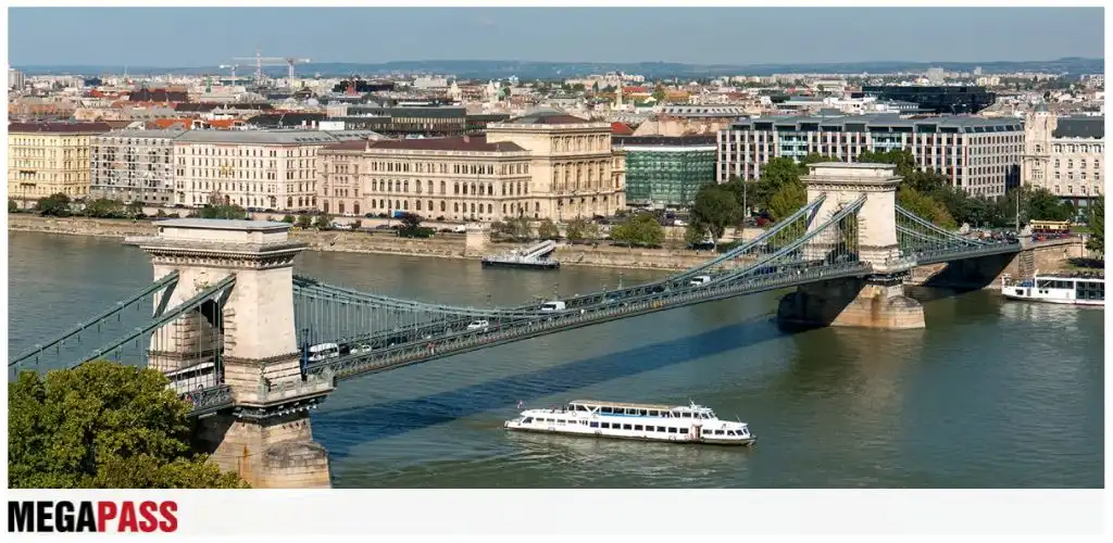 Aerial view of a city with a large suspension bridge over a river and buildings along the shoreline. A boat is sailing on the water.