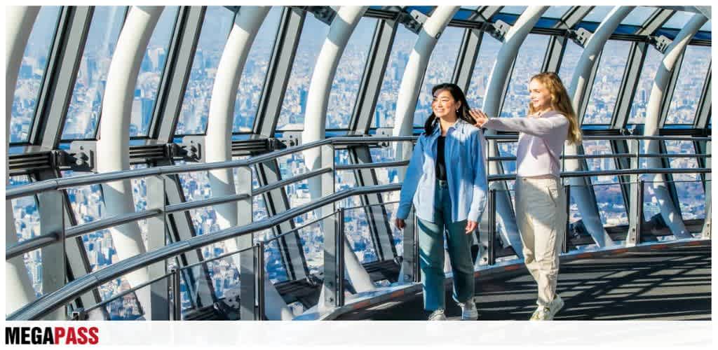 Two women walk and smile inside a glass observation deck with a city view behind them. The scene is bright and modern.