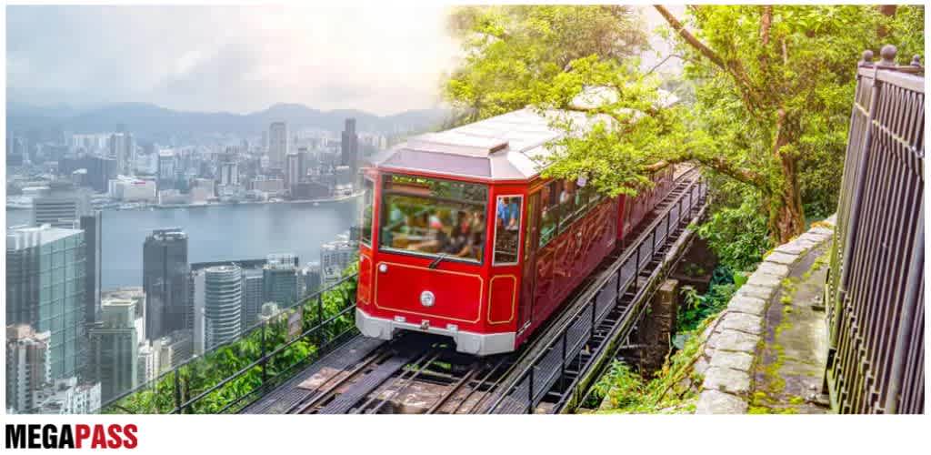 A red cable car traveling along a track with a cityscape and river in the background, surrounded by green trees and a stone path on the side.