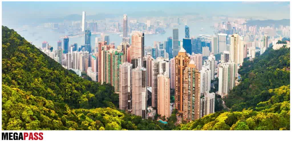 A city skyline with numerous tall buildings surrounded by green hills and mountains.