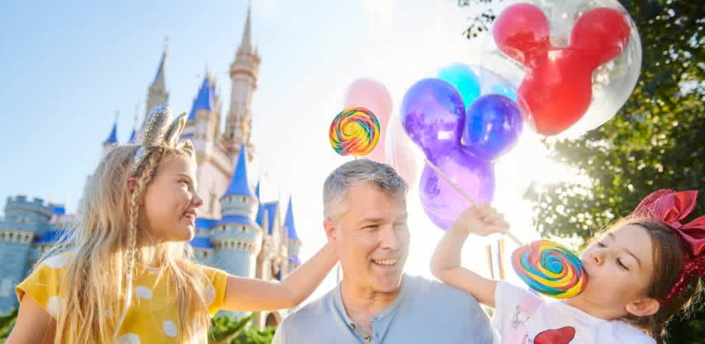 A man enjoying time with two young girls, one holding colorful balloons and a lollipop, in front of a castle at a theme park on a sunny day.