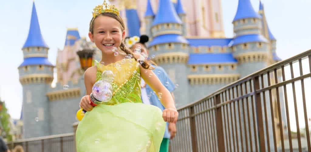 A smiling girl in a yellow and green costume with a crown and bubbles in front of a castle at an amusement park