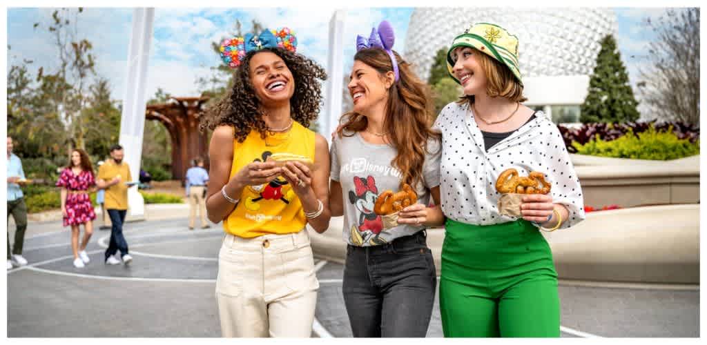 Three women smiling and holding pretzels at an outdoor event with other people in the background