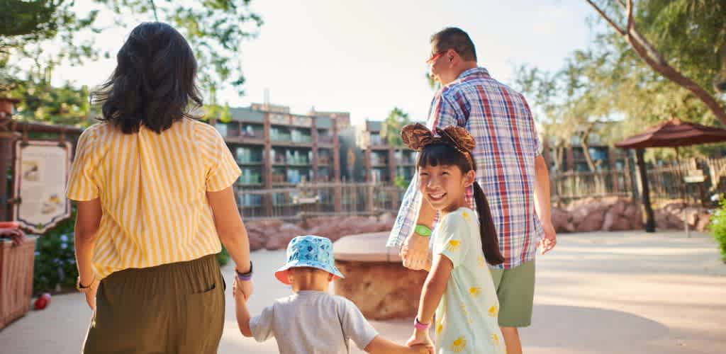 A family of four walking outdoors in a park area with trees and buildings in the background on a sunny day.