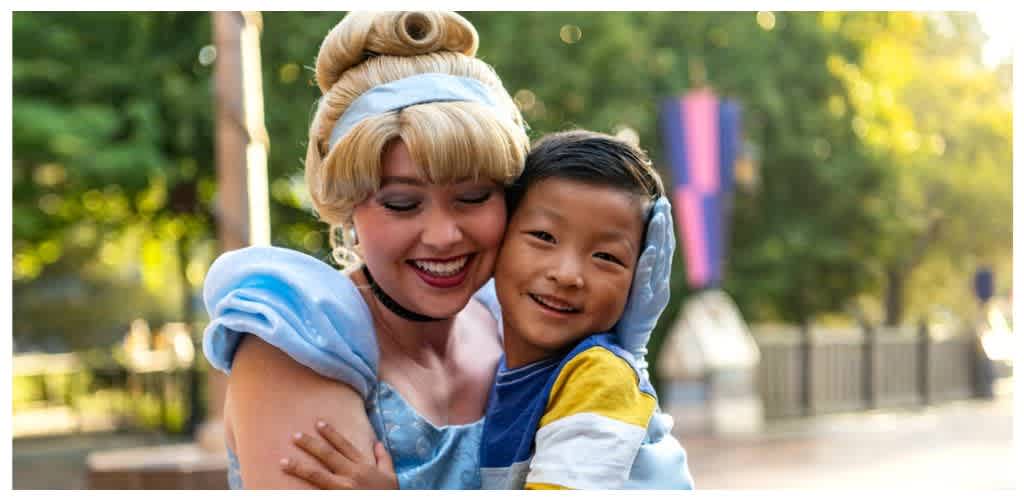 A woman dressed as a fairy tale princess hugs a young boy outdoors with trees and a fence in the background.