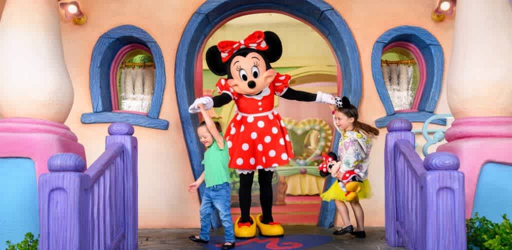 Children are dancing with a person dressed as Minnie Mouse in front of a colorful animated house.