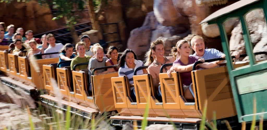 A group of happy people enjoying a roller coaster ride outdoors, smiling and laughing as the train moves through a rocky, themed amusement park area.