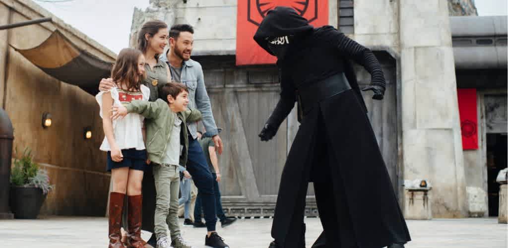 A family watches a person dressed as a character with a hood and cape perform a stunt outdoors in an urban setting.