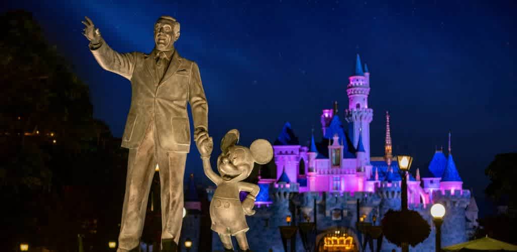 Night view of Walt Disney and Mickey Mouse statues in front of a colorful illuminated castle at Disneyland California.