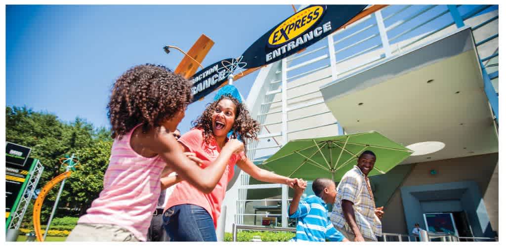 Four people smiling and playing outside an amusement park entrance with a colorful sign and a green umbrella on a sunny day.