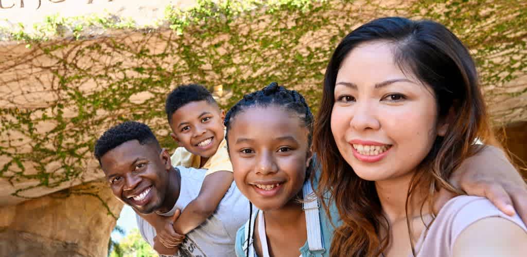 A diverse family of four smiling outdoors in front of a stone wall covered with green vines.