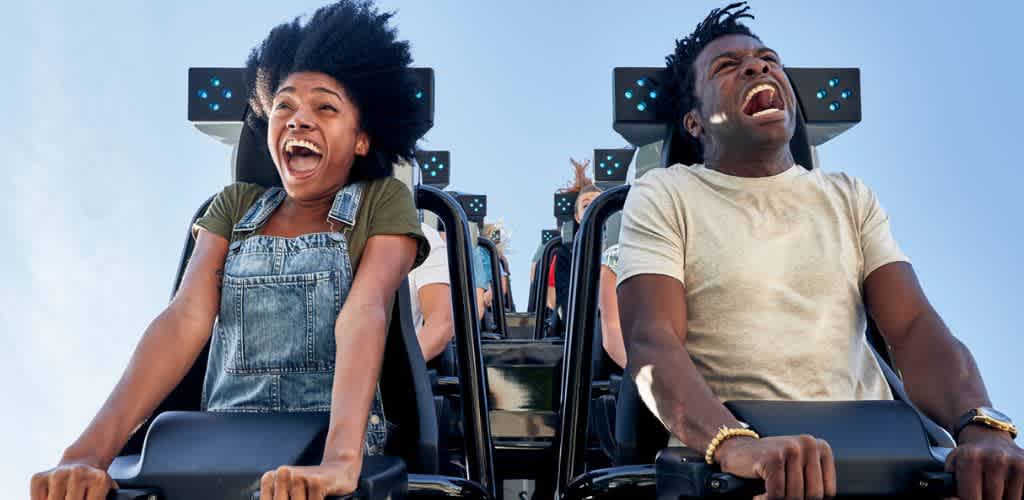 Two people are riding a roller coaster with excited and joyful expressions on their faces under a clear blue sky