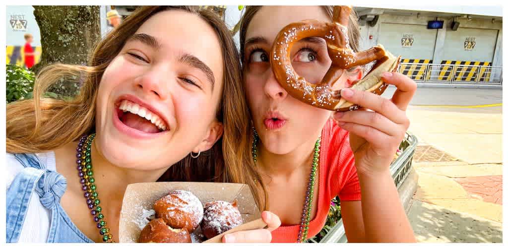 Two young women smiling and enjoying donuts outdoors, wearing colorful beads, with a background of buildings and trees.