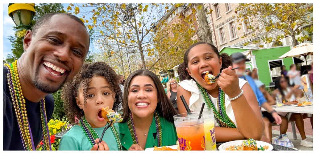 A family of four celebrating Mardi Gras outdoors with colorful beads, eating cake, and enjoying drinks on a festive street with trees and buildings in the background