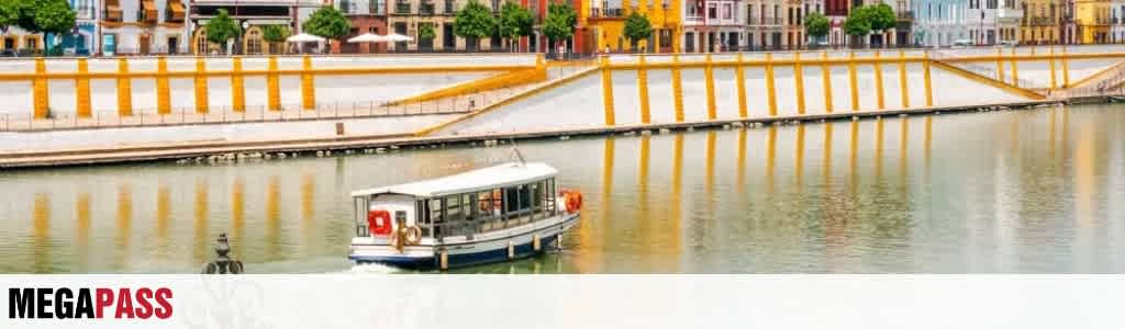 A boat is sailing on a calm waterway near a yellow and orange wall with buildings and trees in the background.