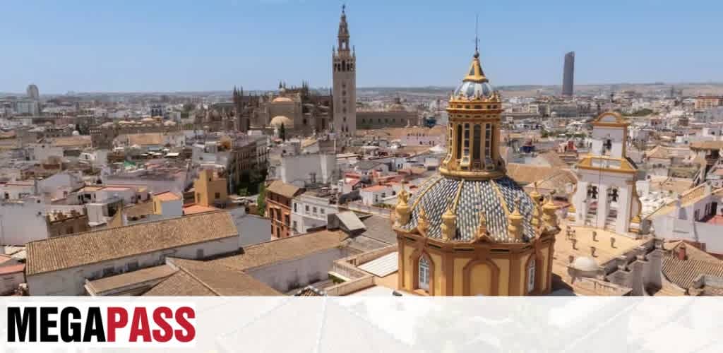 Aerial view of a historic European city with ornate rooftops, a tall clock tower, and a prominent domed church with colorful tiles.