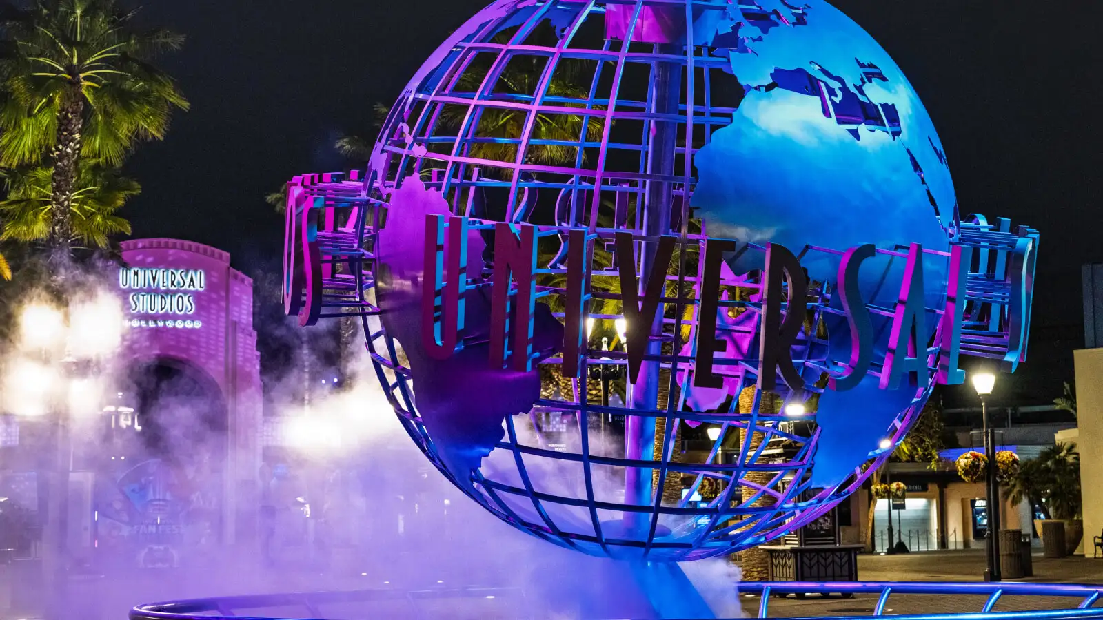 A large illuminated globe sculpture with the Universal Studios logo, surrounded by colorful lights and fog, located outside at night.