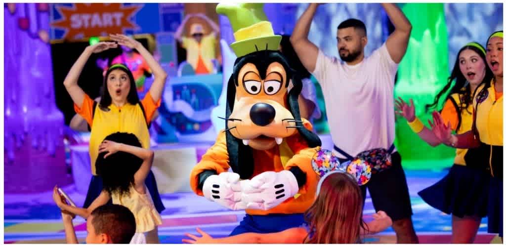 People children and adults celebrating at a colorful indoor party with a life-sized cartoon character mascot in the center, surrounded by excited guests and vibrant decorations
