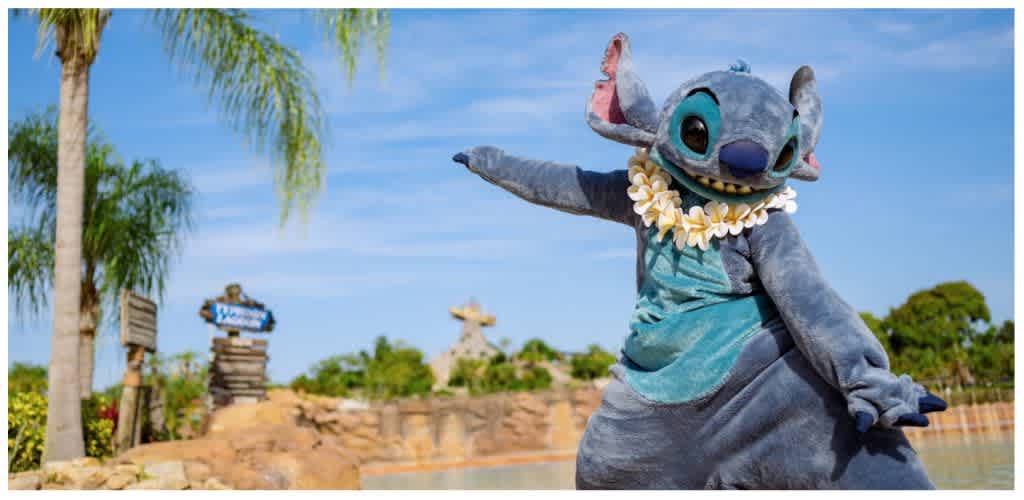 Costumed character of Stitch from Lilo and Stitch standing outdoors with palm trees and a blue sky in the background.