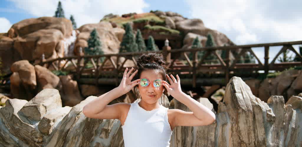 A girl with glasses standing outdoors near rocks and a wooden bridge on a sunny day.