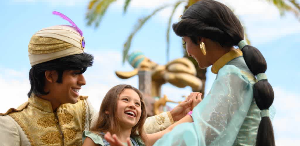 A young girl smiles and laughs as she interacts with two adults dressed in elaborate costumes outdoors during the daytime. The scene appears joyful and festive.