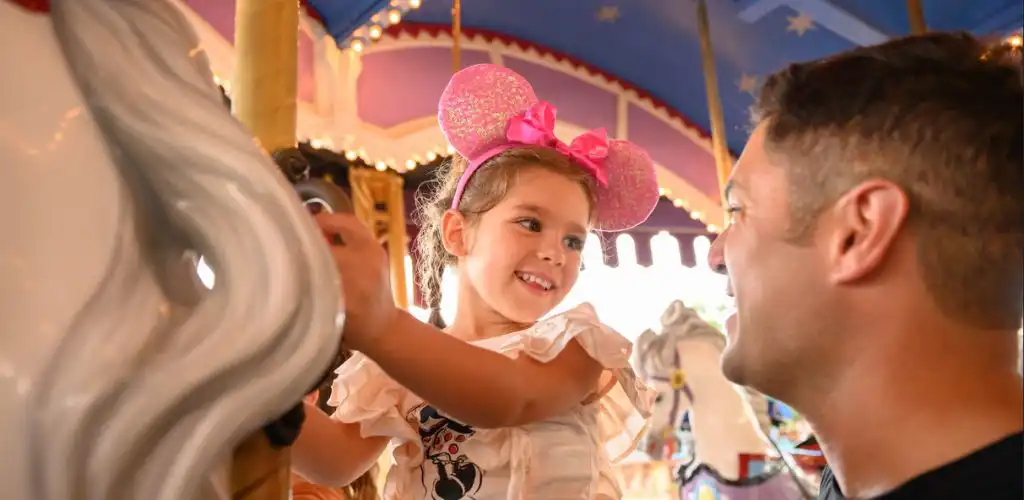 A young girl wearing pink Minnie Mouse ears and a ruffled dress smiles at a man in a festive amusement park setting.