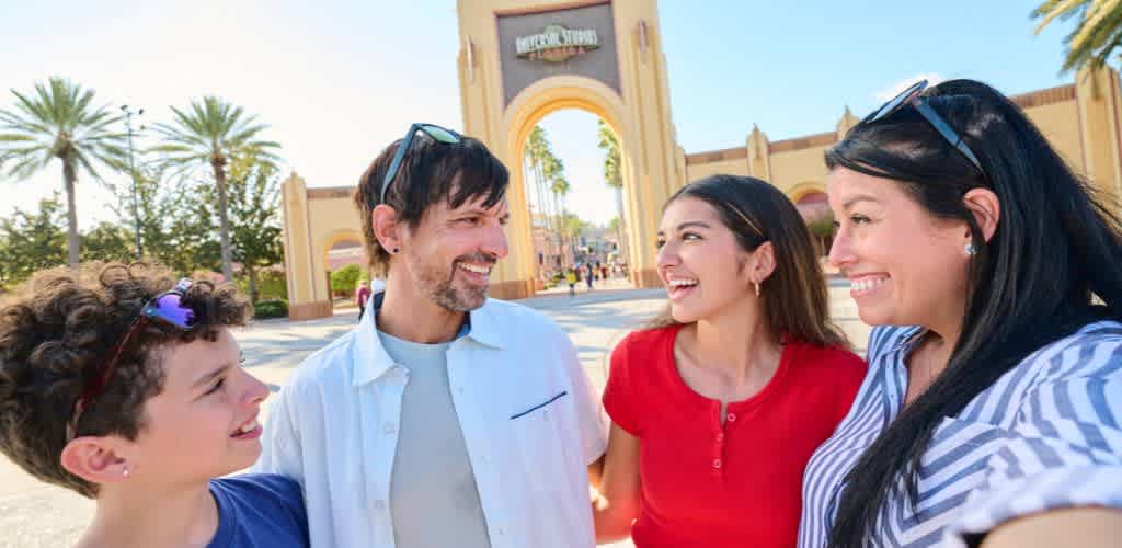 Four smiling people with sunglasses on their heads stand outdoors near an entrance gate with palm trees in the background