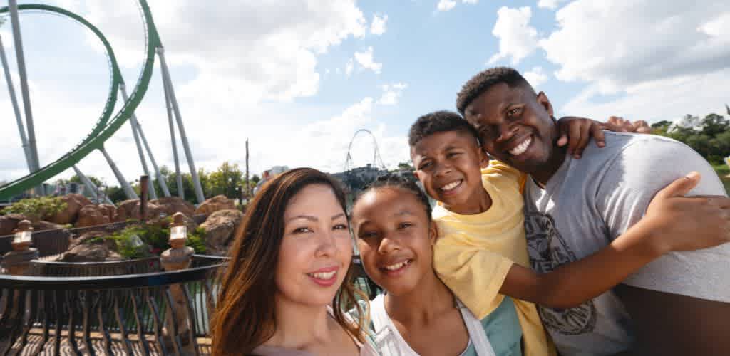 A family of four enjoying a day at an amusement park with roller coasters and clear blue skies behind them.