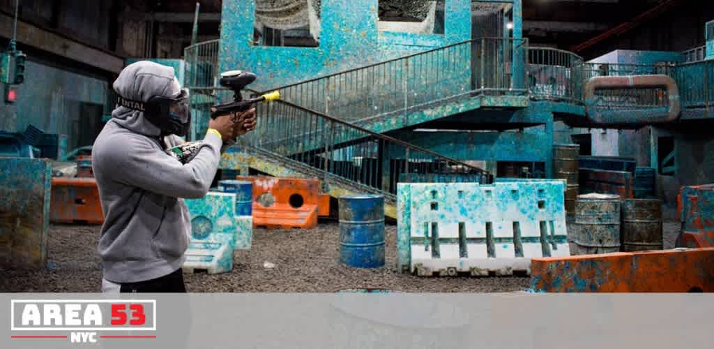 Person dressed in indoor paintball gear holding a paintball gun in an indoor paintball arena with colorful barriers and obstacles