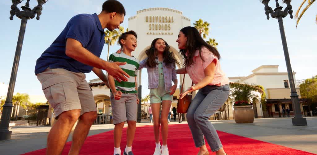 A family with two children and two adults laughing and playing together on a red carpet outside a theme park entrance during daytime