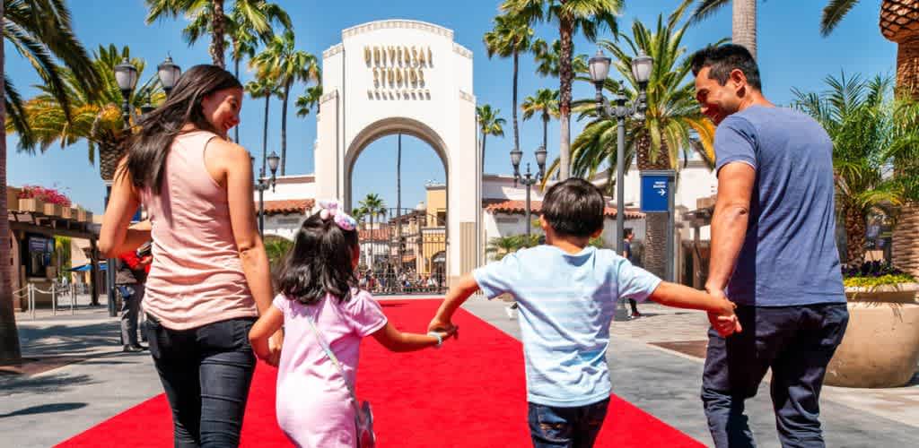 A family walks on a red carpet at an amusement park entrance with palm trees and a clear blue sky behind them.