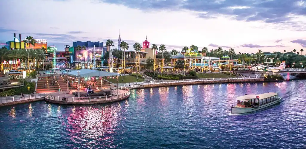 A vibrant waterfront area with colorful buildings, palm trees, and a boat on the water at dusk with city lights reflecting on the surface