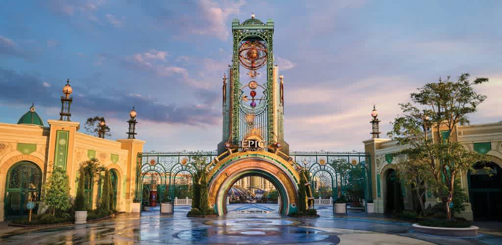 The image shows a colorful amusement park entrance with a clock tower, decorative arches, and lush greenery under a partly cloudy sky.