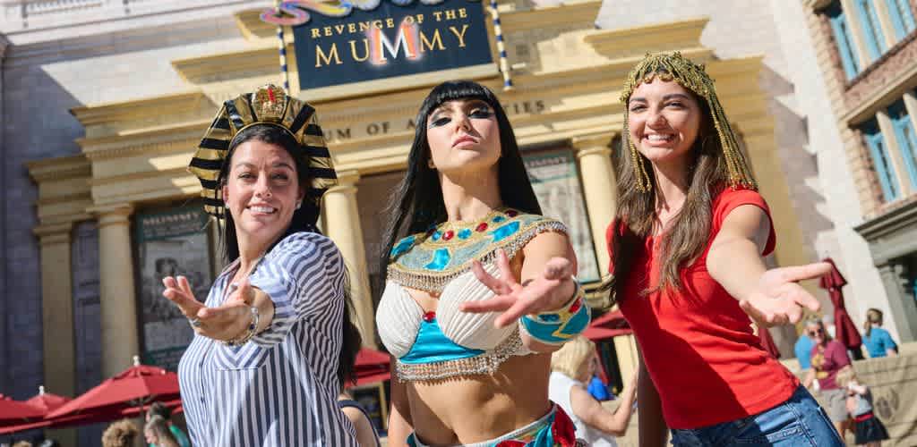 Three women dressed as ancient Egyptian characters perform in front of a movie theater during a themed event.