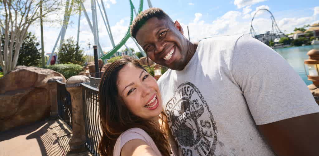 A smiling couple taking a selfie outdoors at an amusement park with clear skies and a lake in the background.