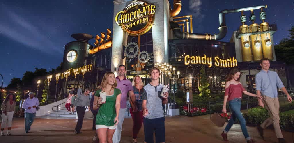 People walking in front of a themed chocolate factory building at night with signs for chocolate and candy shop.