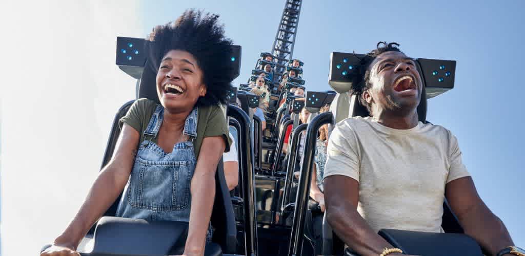 Two people are enjoying a roller coaster ride with big smiles and open mouths on a sunny day.