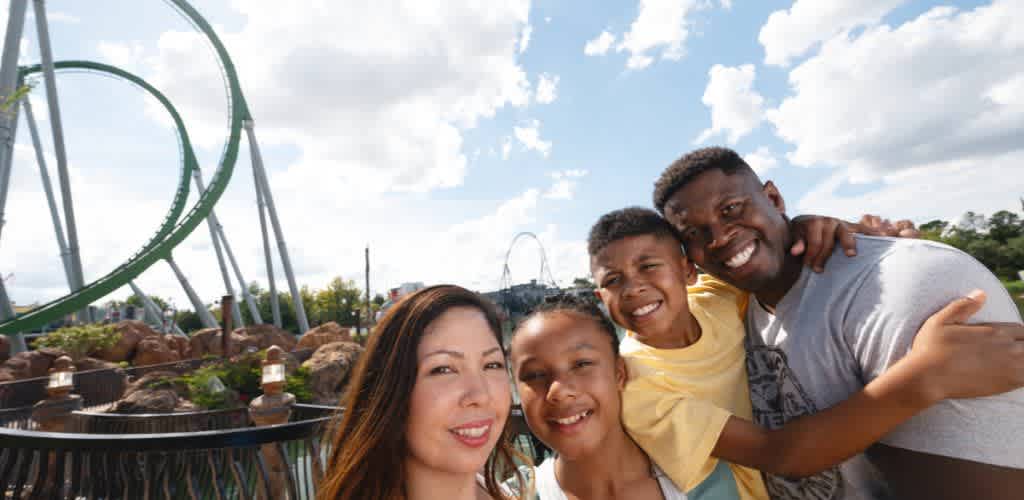 A happy family of four smiling and hugging each other outdoors at an amusement park with a roller coaster in the background