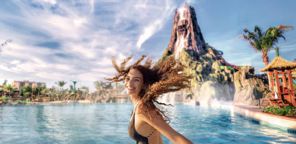 A smiling woman with curly hair enjoying a tropical water park with a volcano and palm trees in the background during a sunny day.