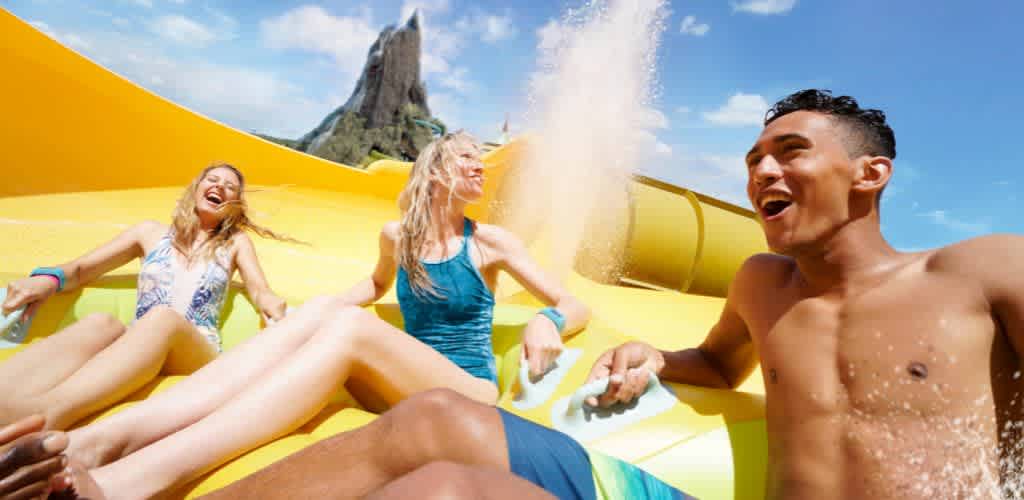 Three people are enjoying a water slide outdoors on a sunny day, laughing and having fun on a bright yellow slide with a mountain in the background.