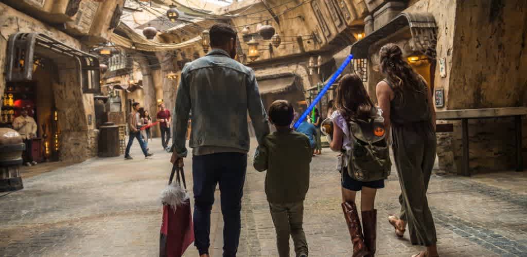 A family walks together through a themed indoor cavernous space with stone walls and warm lighting, carrying shopping bags and a toy lightsaber.