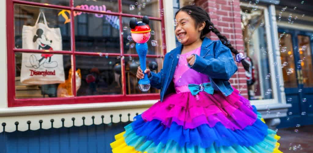 A young girl in a colorful tutu dress laughs and plays with bubbles outside a storefront with a Disney store sign and window display.
