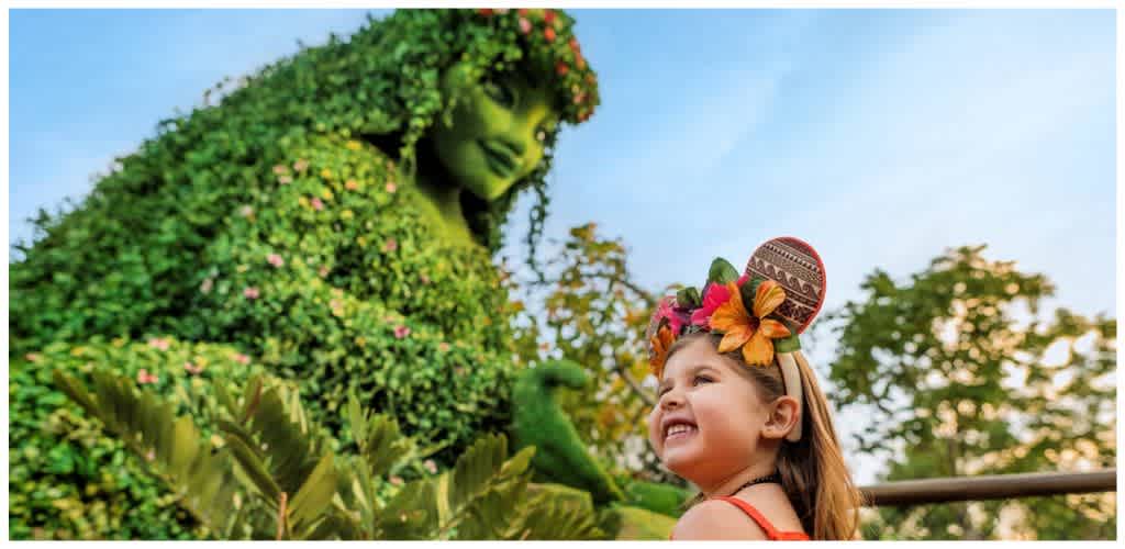 A young girl in colorful festive ears smiles up at a large, lush green topiary sculpture of a woman with flowing hair.