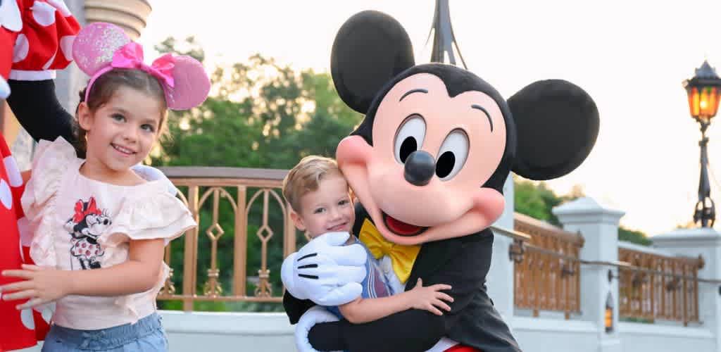 Two children hugging a person dressed as Mickey Mouse outdoors at a theme park during sunset.