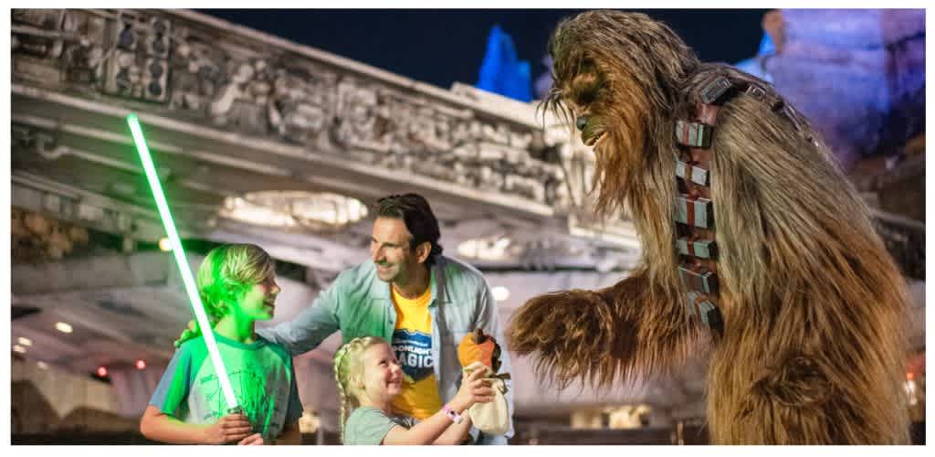 A family and a person in a Chewbacca costume enjoy a themed event with children holding a green lightsaber and a plush toy, smiling indoors.